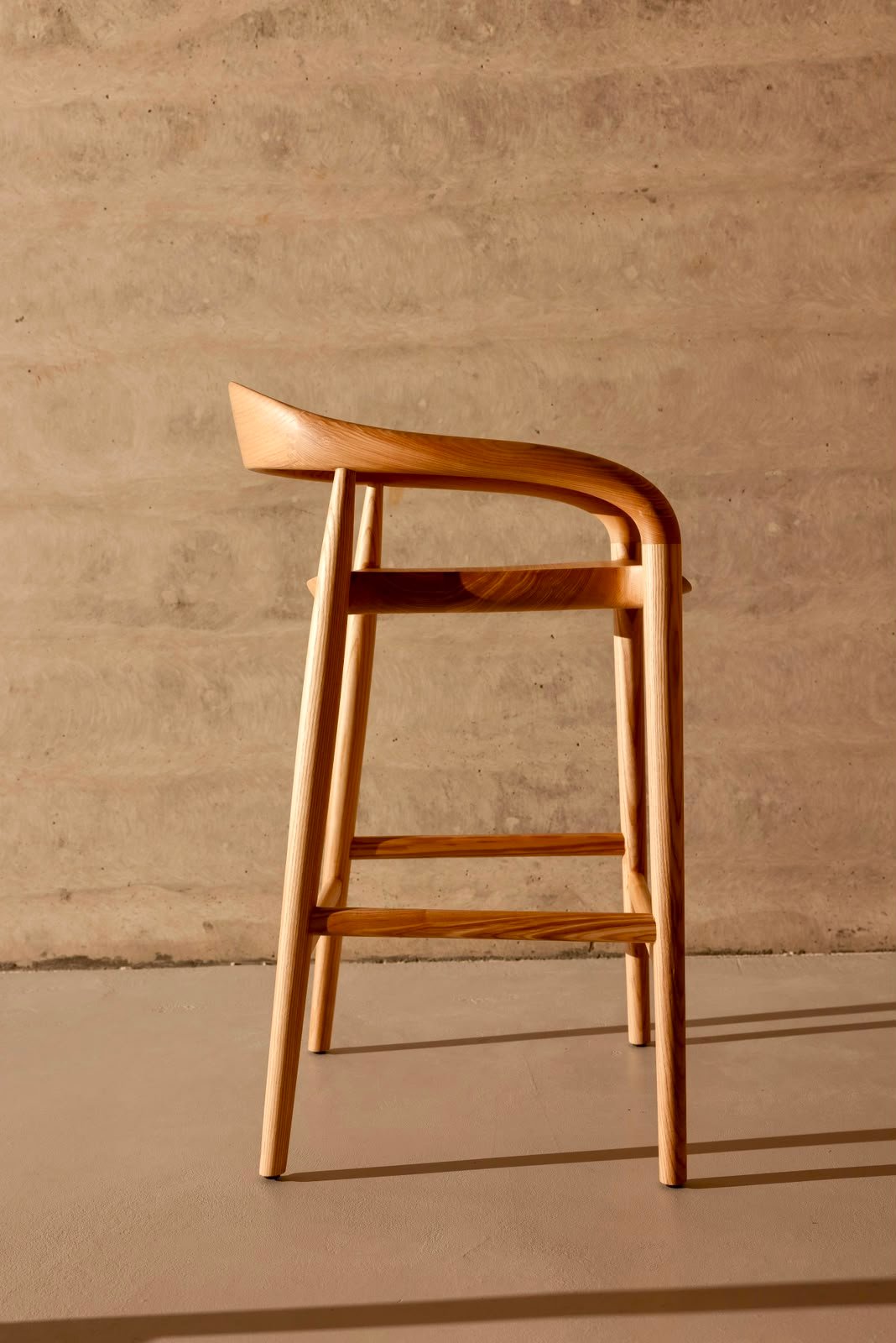 Side profile of a solid ash barstool showing its curved backrest, tapered legs and footrail